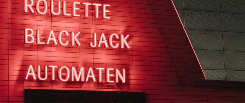 Red neon letters are mounted on a dark tiled wall, displaying the words Poker, Sic Bo, Roulette, Black Jack, and Automaten.