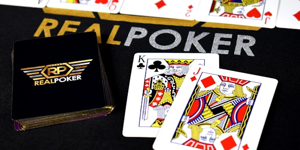 Poker-themed setup featuring a deck of cards on a table with the words 'REAL POKER' visible. Two cards, the King of Clubs and Jack of Diamonds, are prominently displayed. The design is vibrant, with detailed illustrations on the cards.