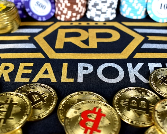Stacks of poker chips in various colors are arranged in the background, while in the foreground, there are numerous golden coins featuring the Bitcoin symbol. The text 'REALPOKER' is prominently displayed in gold and silver lettering on a dark background.