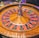 A roulette wheel with alternating red and black numbered pockets on a wooden platform. The wheel contains a small white ball resting in one of the pockets, indicating gameplay. Surrounding the wheel are stacks of colorful poker chips, suggesting a casino environment.