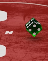 Two green dice with rounded edges are positioned on a red surface that seems to be part of a game board. The surface has white text and numbers on it, including the word 'LUCKYTOPSPIN' in capital letters.