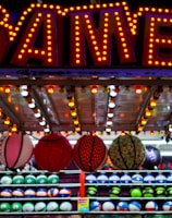 A brightly lit carnival game stall with a large 'GAMES' sign at the top surrounded by red and yellow lights. Various sports balls, including basketballs and soccer balls, are hanging from the ceiling and neatly arranged on shelves below.