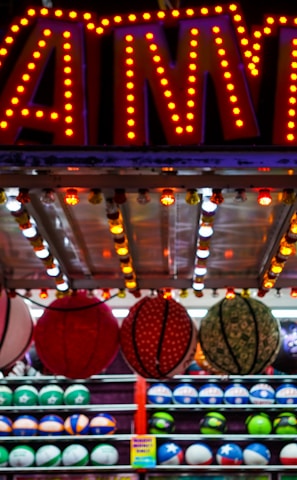 A brightly lit carnival game stall with a large 'GAMES' sign at the top surrounded by red and yellow lights. Various sports balls, including basketballs and soccer balls, are hanging from the ceiling and neatly arranged on shelves below.